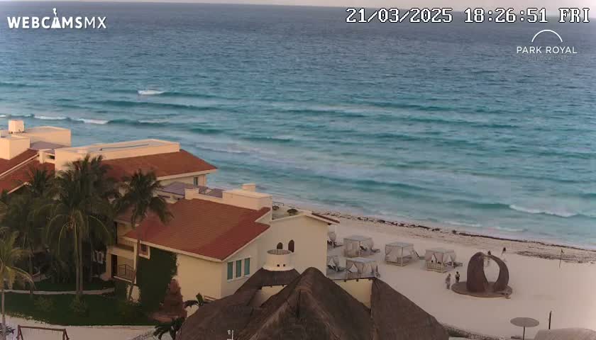 A beachfront resort with reddish-brown roofs and palm trees overlooks a calm, turquoise ocean under a clear evening sky.