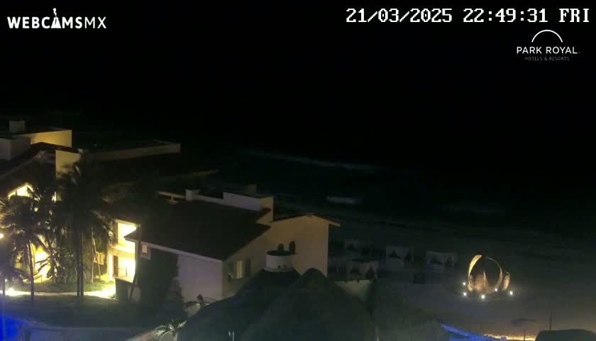 A nighttime view of a beachfront resort with palm trees and gently lapping waves.