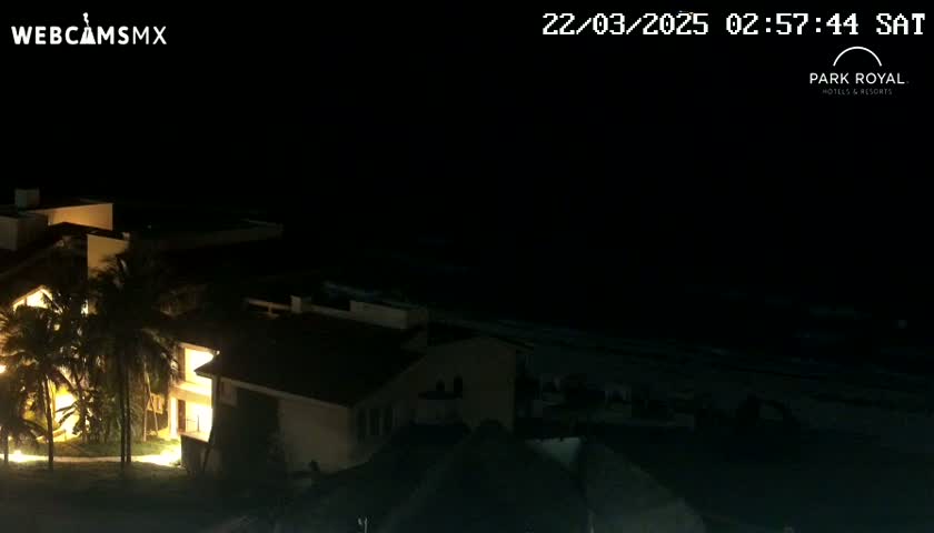 A nighttime view of a coastal resort complex under a dark sky shows illuminated buildings and palm trees near a dark ocean.