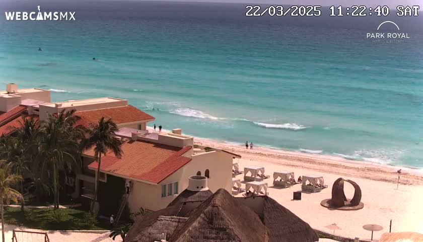 A beachfront resort with red-tiled roofs overlooks a sandy beach and clear turquoise ocean under a sunny sky.