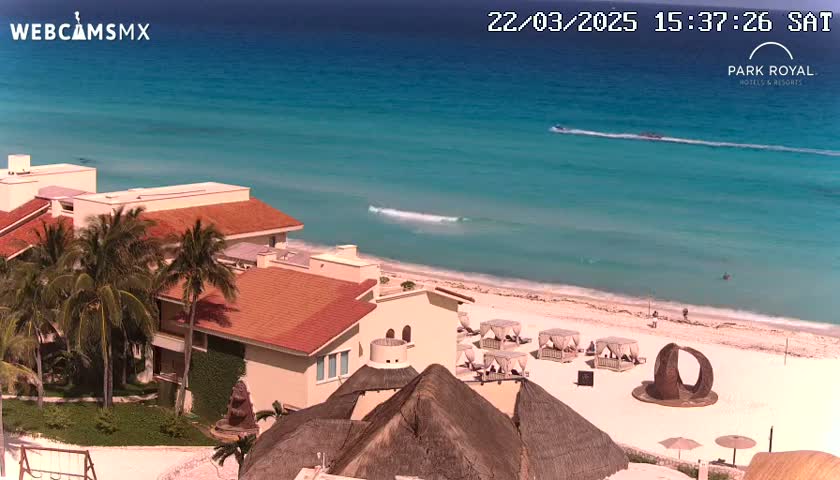 An aerial view shows a resort with red-tiled roofs near a beach with turquoise water under a sunny sky.