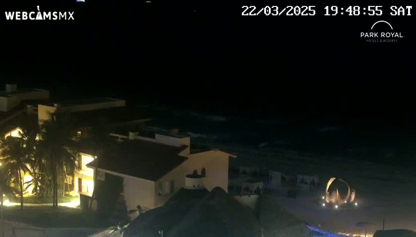 At night, illuminated buildings sit near a dark ocean with gentle waves under a clear sky.