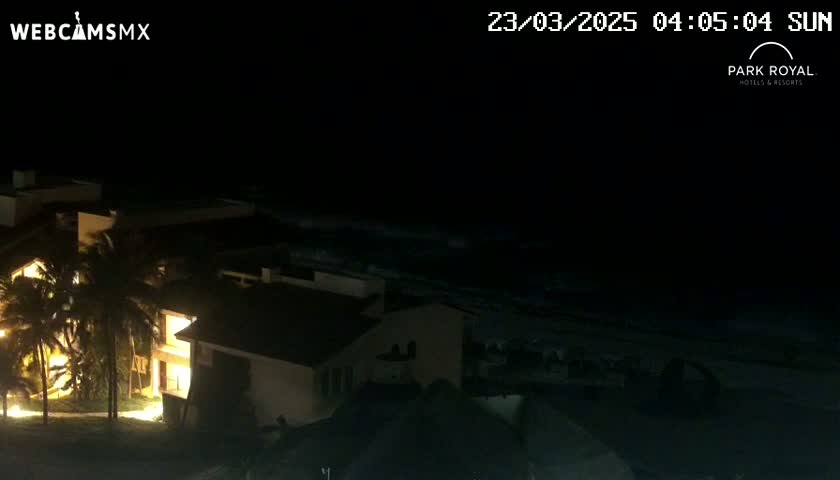 At night, illuminated buildings are situated near a dark ocean under a clear sky.