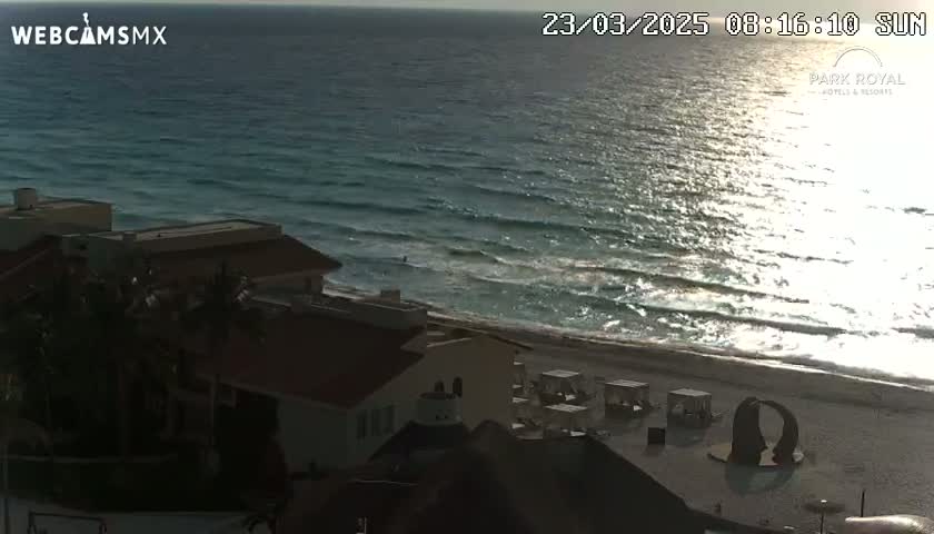 A beach scene shows a partly sunny day with ocean waves lapping a sandy shore near buildings and outdoor furniture.