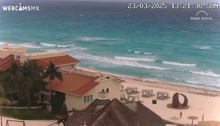 A beachfront resort with red-tiled roofs overlooks a turquoise ocean with moderate waves under a somewhat cloudy sky.