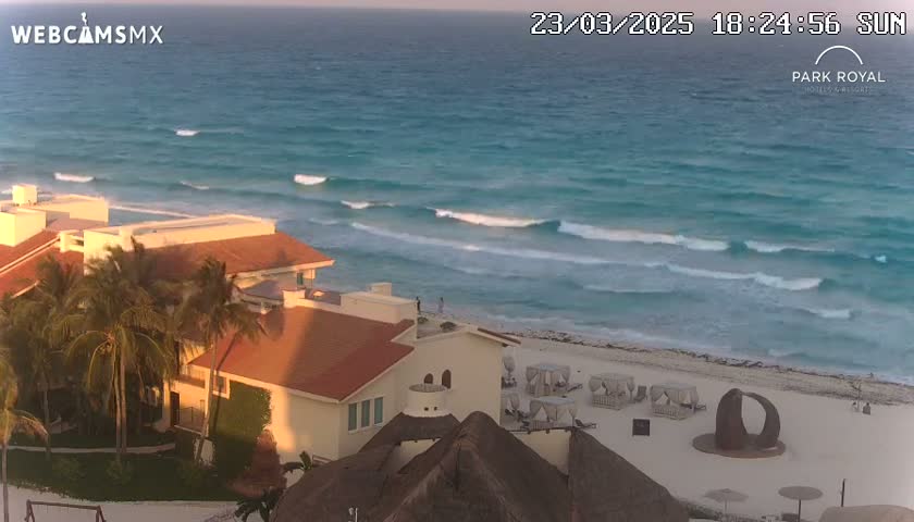 A coastal resort building with palm trees is situated near a beach with gentle waves under a sunny sky.