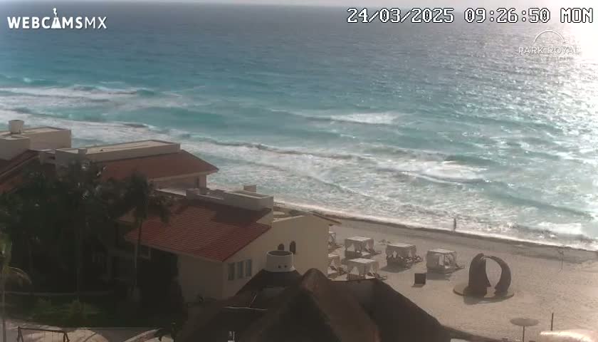An ocean with moderate waves laps a sandy beach where white beach umbrellas are set up near a group of low-rise buildings under a mostly sunny sky.