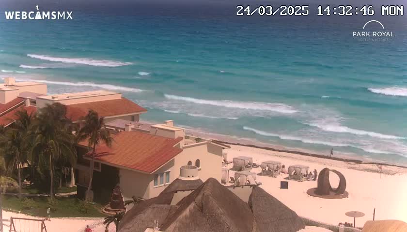 An ocean with waves lapping a sandy beach, in front of a resort with red-tiled roofs and palm trees, under a mostly clear sky.