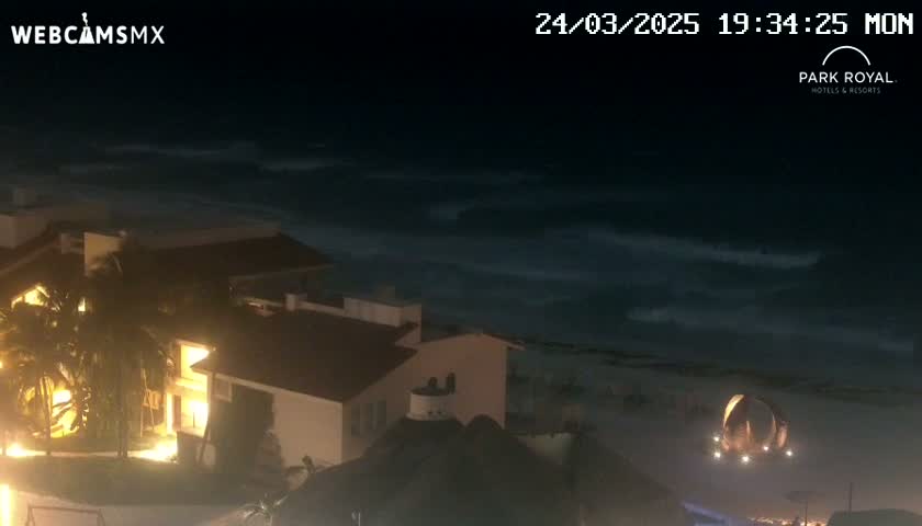 At night, a beach with moderately rough waves is seen from above a brightly lit building complex.