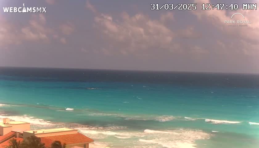A partly cloudy day shows a turquoise ocean with waves breaking on a sandy beach near buildings with red tile roofs.
