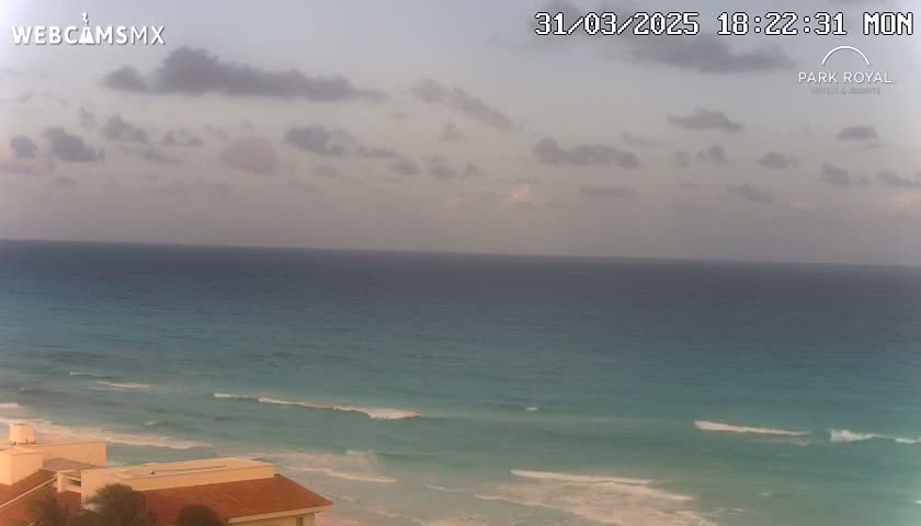 A partly cloudy evening sky overlooks a calm turquoise ocean with small waves lapping a sandy shore, partially visible buildings in the foreground.