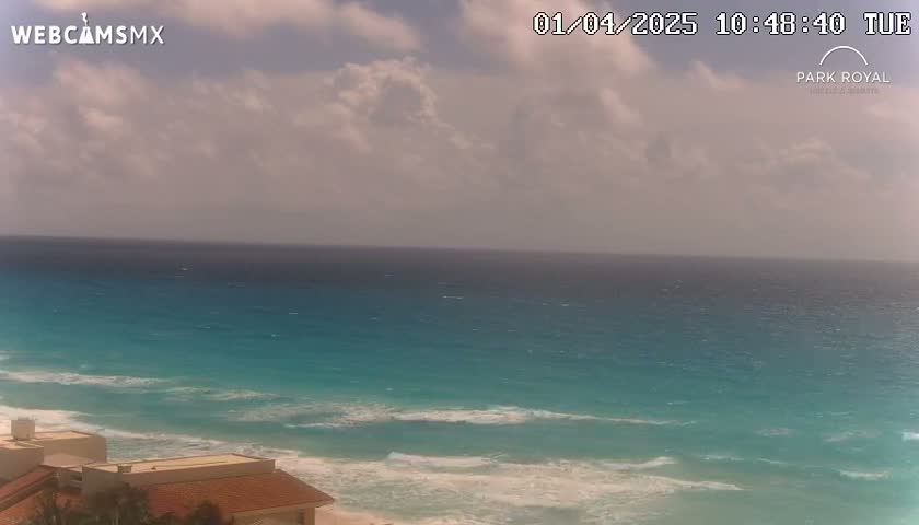 A partly cloudy day shows a view of a turquoise ocean with waves lapping a sandy beach near some buildings.
