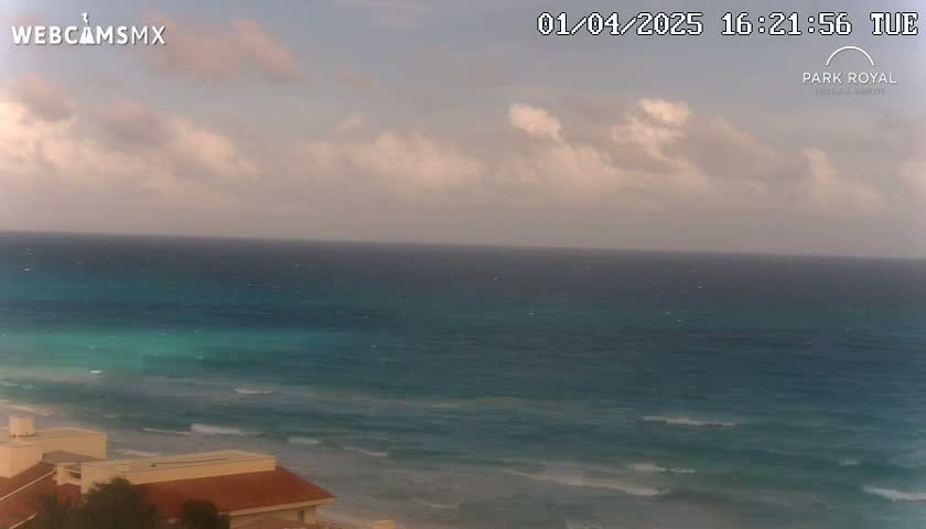 A partly cloudy sky overlooks a teal ocean with small waves lapping a sandy beach, and a portion of a building is visible in the bottom left corner.