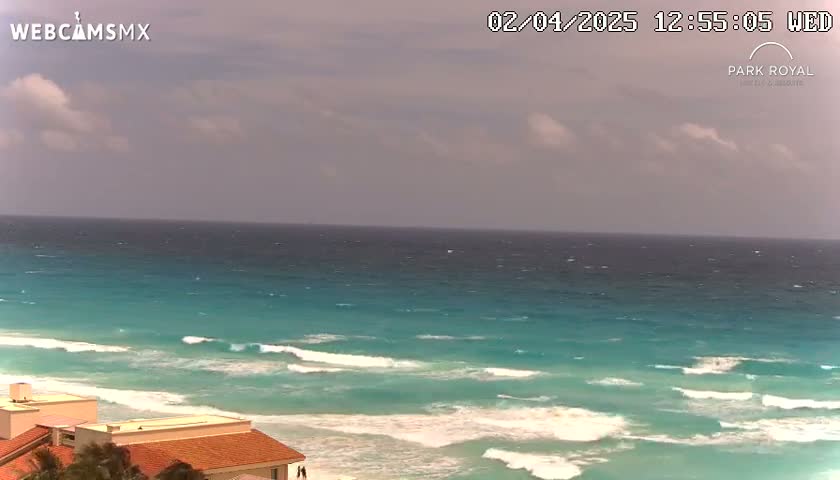 A partly cloudy sky overlooks a teal ocean with waves breaking on a sandy beach near low-lying buildings.