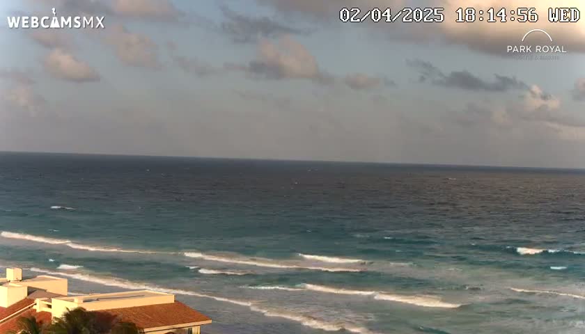 A partly cloudy sky overlooks a teal ocean with small waves lapping a sandy shore, where a portion of a building is visible in the bottom left corner.