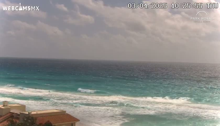 A partly cloudy sky overlooks a turquoise ocean with white waves breaking on a sandy beach, near a building with a red tile roof.
