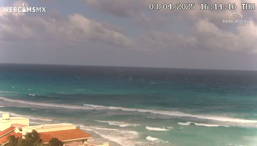 An ocean scene with partly cloudy skies shows a beach with waves rolling onto the shore near buildings with red tile roofs.