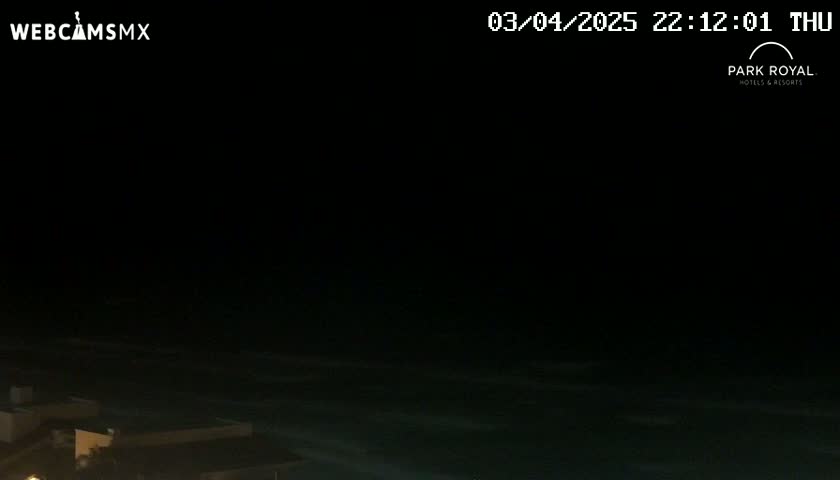 A nighttime view of dark ocean waves gently lapping the shore near some low-lying buildings.