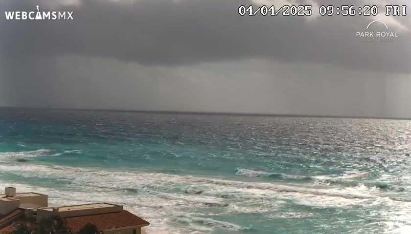 A partly cloudy day shows an ocean with waves crashing on a sandy beach near buildings.