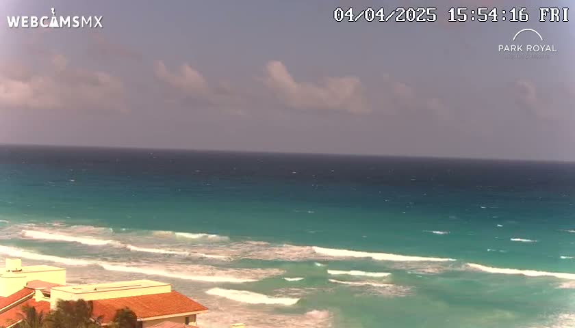 A partly cloudy sky overlooks a turquoise ocean with small waves breaking on a sandy shore near low-lying buildings.