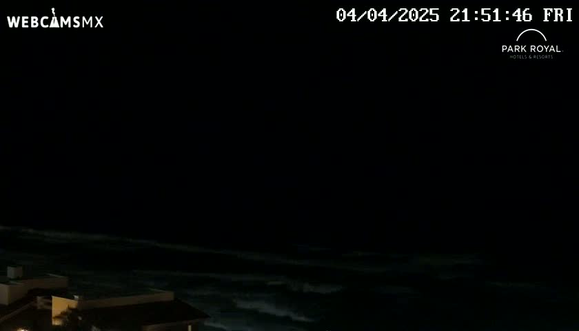 A nighttime, dark view of a wavy ocean and the roofs of buildings under a black sky.