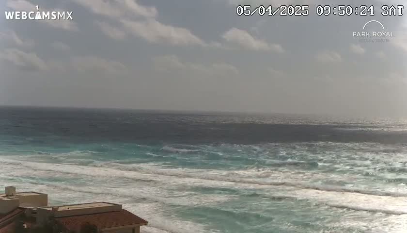 A partly cloudy day shows a view of turquoise ocean waves breaking on a sandy shore near a building.