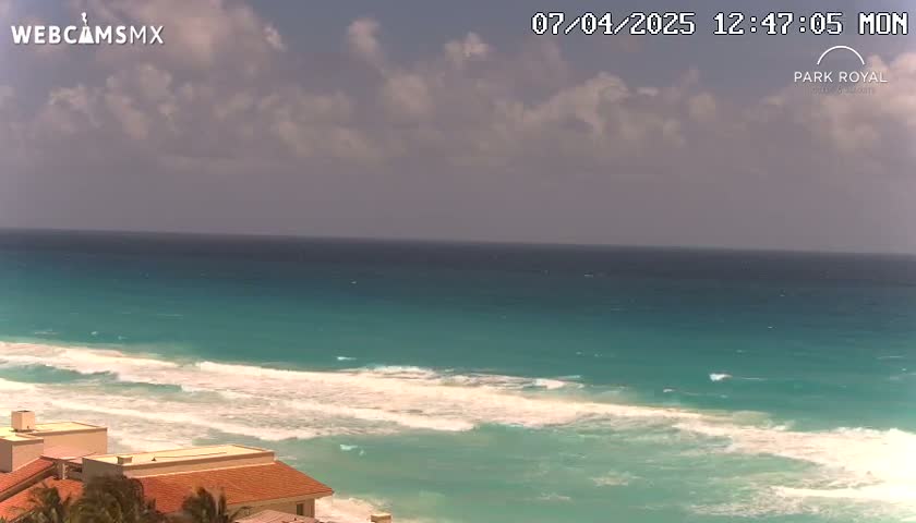 A partly cloudy sky overlooks turquoise ocean waves breaking on a beach near low-rise buildings.