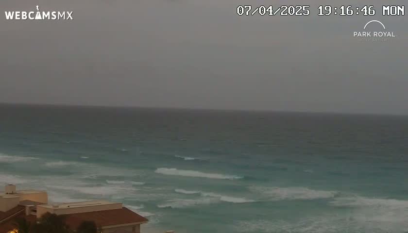 A dark gray overcast sky hangs over a teal ocean with small waves breaking near a beach and several buildings visible on the shore.