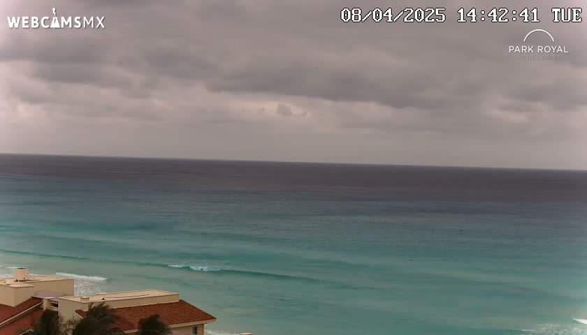 An overcast sky hangs over a teal ocean with small waves lapping a sandy beach, where a portion of a building is visible in the bottom left corner.