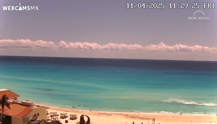 A mostly sunny day shows a panoramic view of a beach with turquoise water, a sandy shore, some buildings, and a few people.