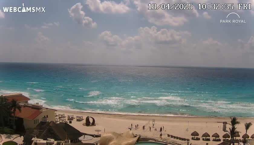 A partly cloudy day shows turquoise water lapping a sandy beach with resort buildings and a few people visible.