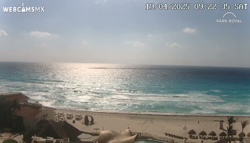 A partly sunny day shows turquoise ocean waves lapping a sandy beach in front of a resort.