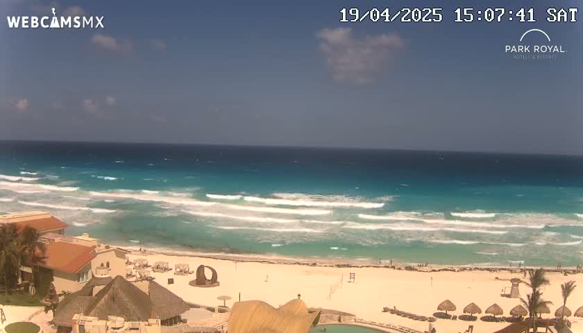 A beach with turquoise water and small waves under a mostly sunny sky, with resort buildings and palm trees visible along the shore.