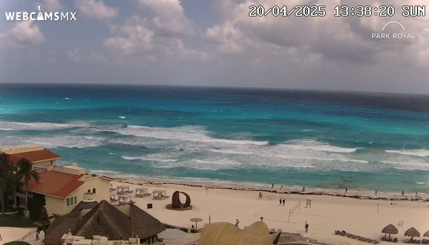 An elevated view shows a beach with turquoise water, small waves, and a few people, under a partly cloudy sky.