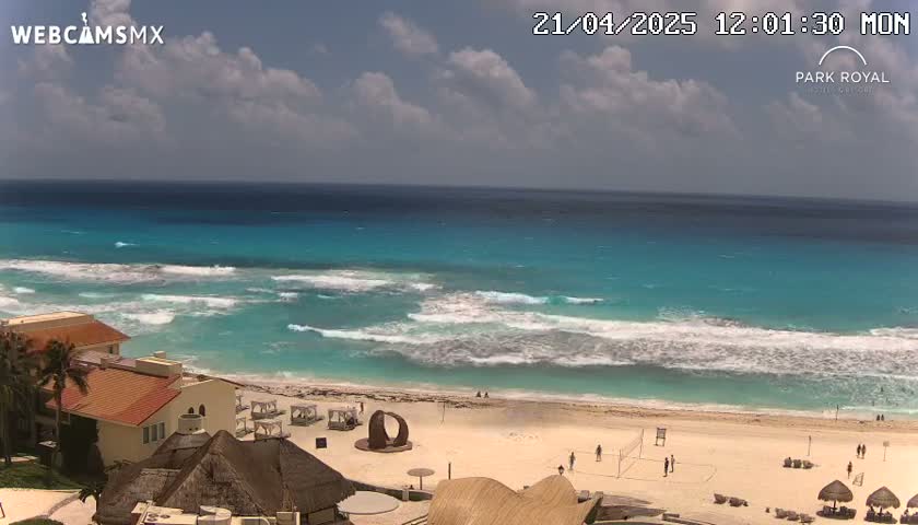 A beach with turquoise water and moderate waves under a partly cloudy sky shows a resort with buildings and beach umbrellas.