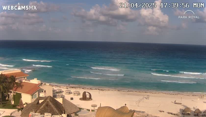 A partly cloudy day shows a beach with turquoise water, gentle waves, and buildings along the shore.