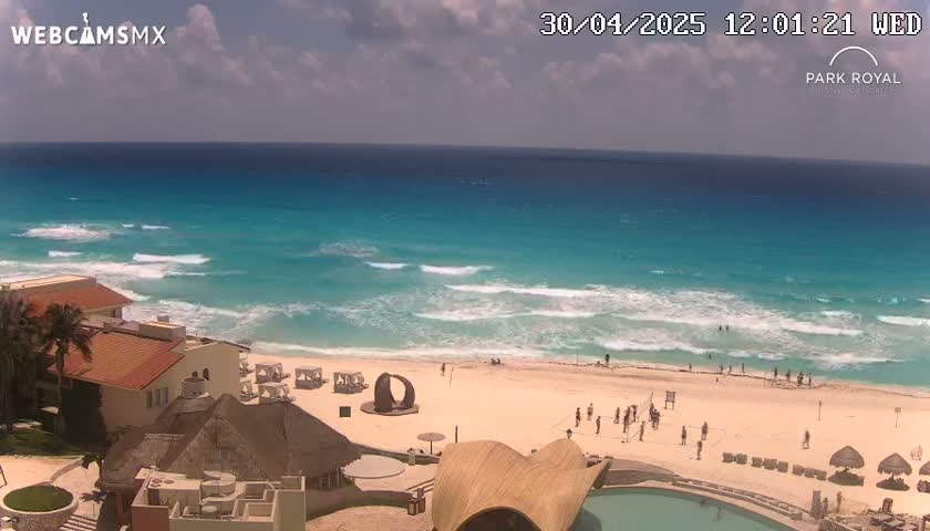 A beach with turquoise water and small waves under a partly cloudy sky is viewed from above, showing buildings and people on the sand.