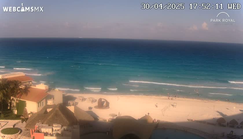 A partly sunny view of a beach with turquoise water and small waves, showing buildings and beachgoers from a high vantage point.