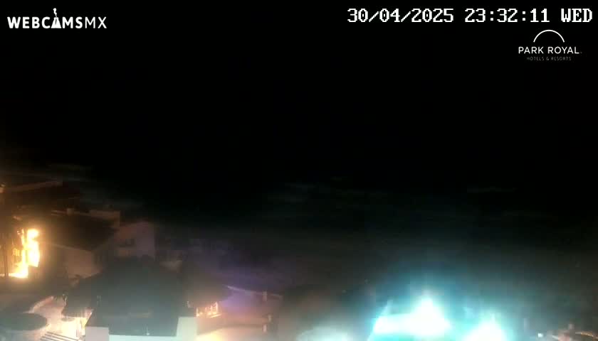 A nighttime view of a coastline shows dark water with a bright blue light source in the lower right, and buildings with lights are visible on the land to the left.