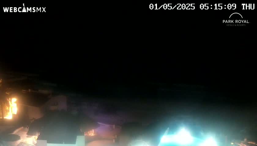 A nighttime, low-angle view shows a dimly lit coastal area with buildings and a bright light source in the lower right corner.