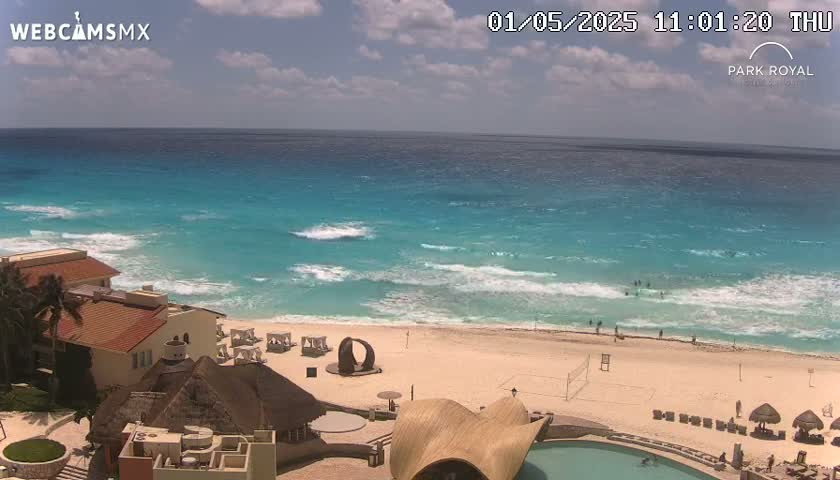 A partly sunny day shows a beach with turquoise water,  light waves, and several buildings overlooking the ocean, with some people visible on the beach and in the water.