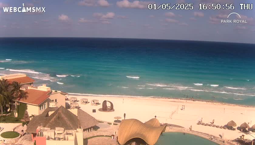 A resort overlooks a beach with turquoise water and gentle waves under a mostly sunny sky.