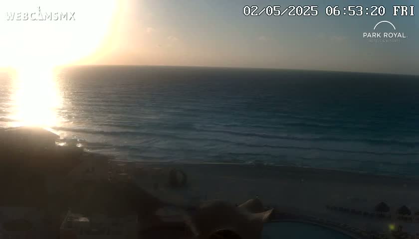 A sunrise over a calm ocean with a sandy beach and resort buildings in the foreground.