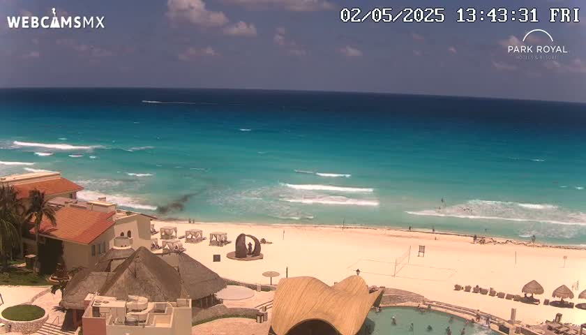 A beach resort with turquoise water and a partly cloudy sky overlooks a sandy beach with small waves.