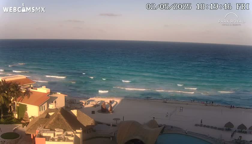 A tranquil ocean scene at sunset shows a beach with several buildings and a few people, under a clear sky.