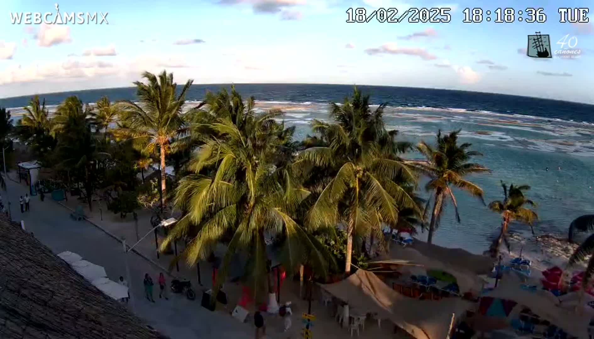 A seaside town with palm trees, a sandy beach, and calm ocean under a partly cloudy sky at sunset.