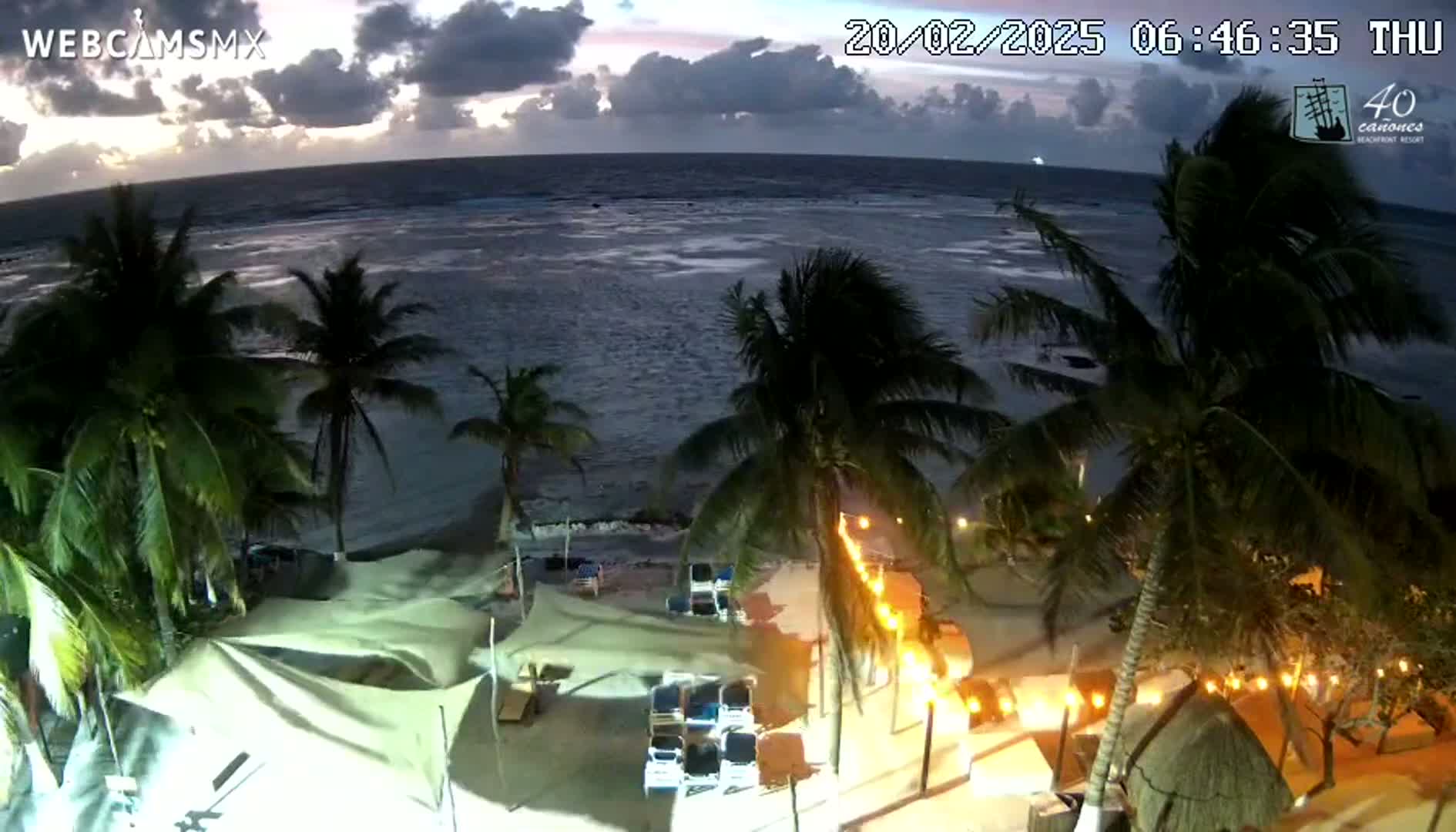 An overhead view shows a beach at twilight with palm trees, beach umbrellas, and string lights, under a partly cloudy sky.