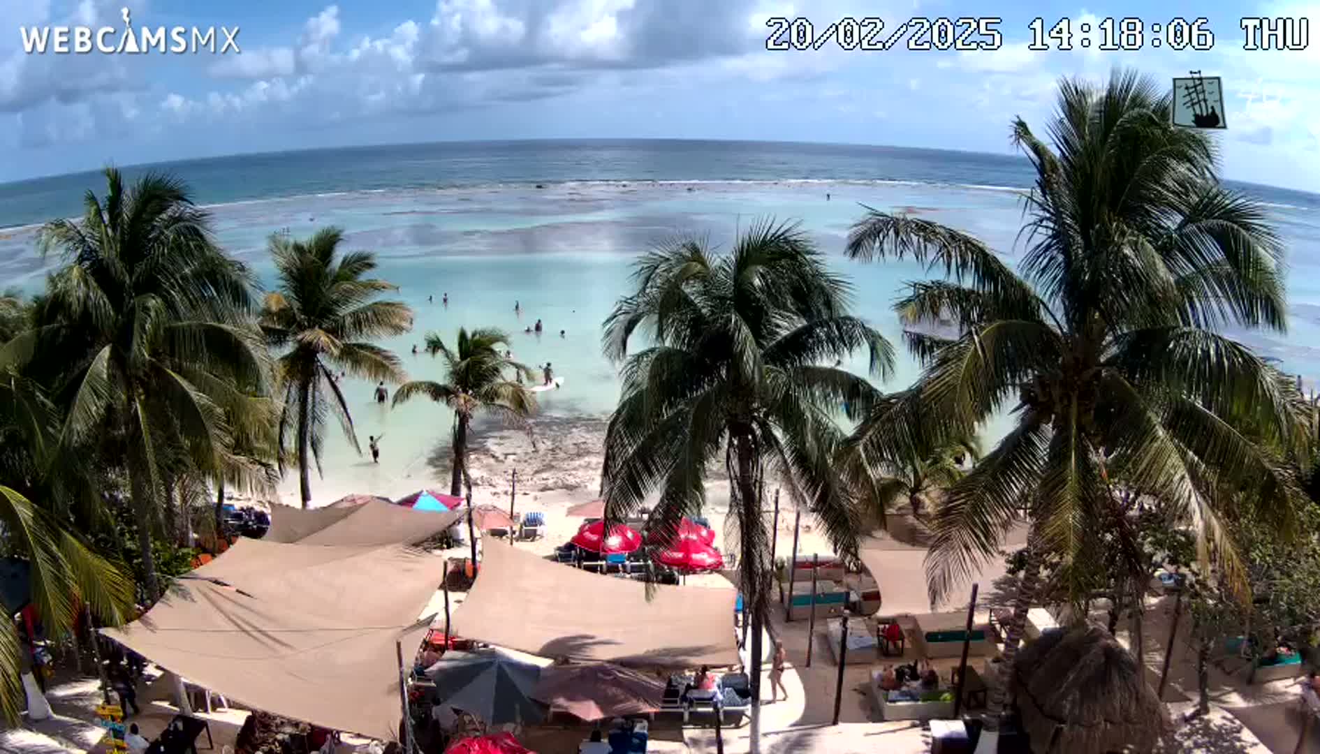 A beach scene on a partly sunny day shows a calm ocean with shallow, light-blue water, a sandy shore with palm trees, and beach umbrellas shading outdoor seating areas.
