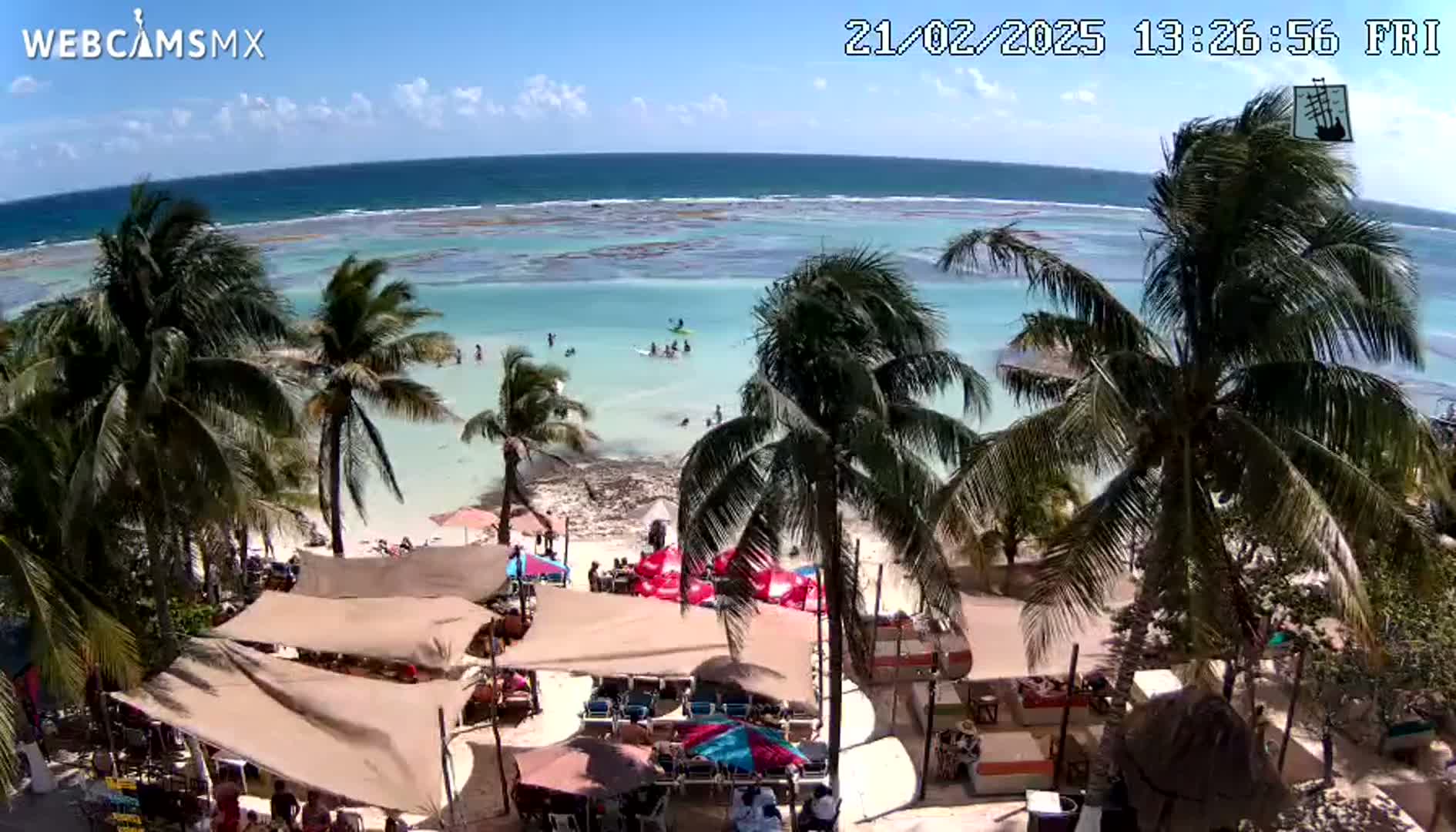 An overhead view shows a beach bar and restaurant with palm trees under a mostly sunny sky, overlooking a calm, shallow turquoise ocean with several people swimming and enjoying the water.