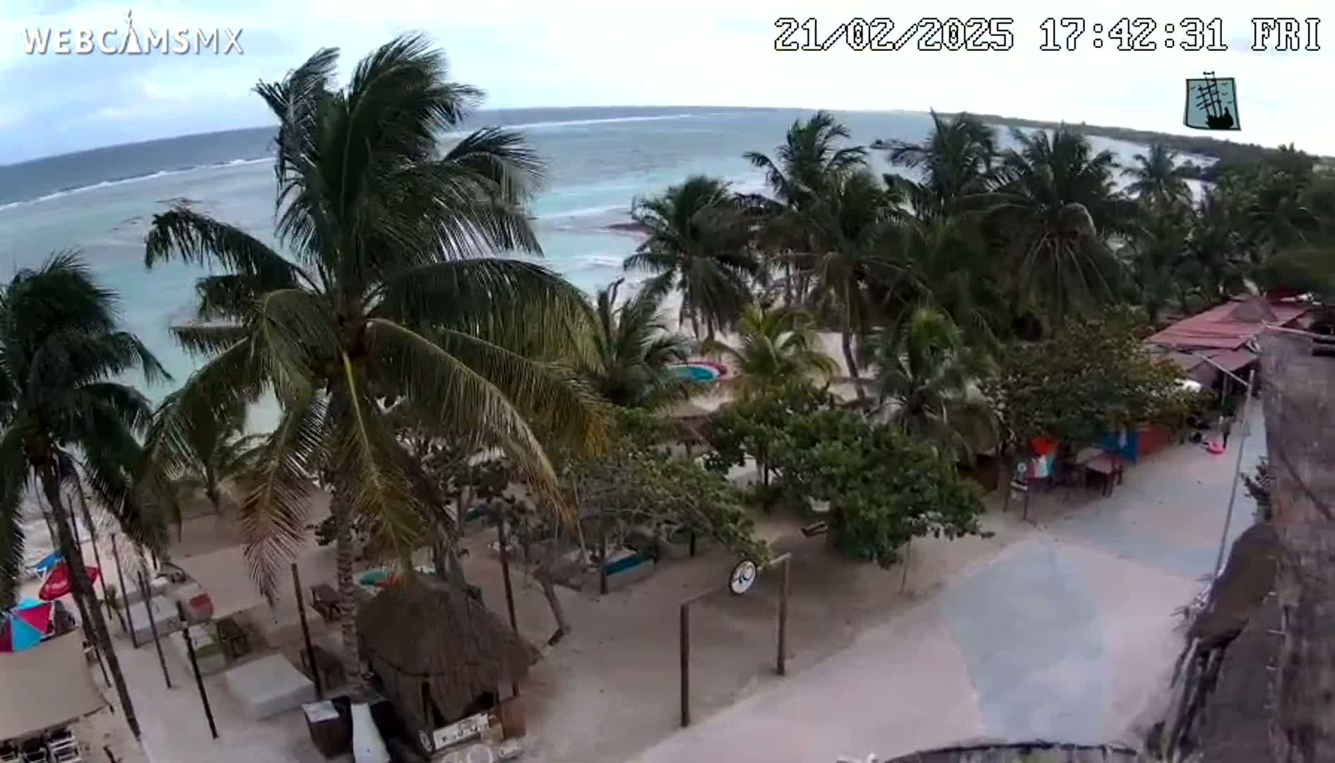 An overhead view shows a beachside area with palm trees, outdoor seating, and small buildings under a partly cloudy sky.